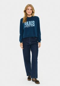 Giovane donna che indossa una felpa blu con la scritta "Paris Athletic Dept.", jeans dritti blu scuro e mocassini marroni, in piedi contro uno sfondo bianco.