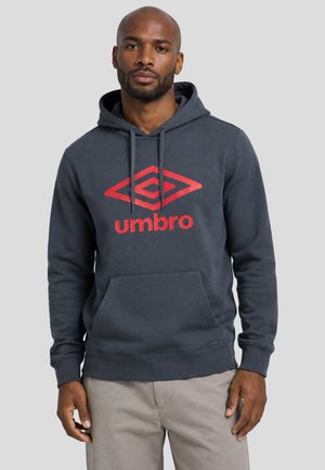 Mann trägt einen dunkelgrauen Hoodie mit rotem Umbro-Logo und hellen Hosen und steht mit neutralem Gesichtsausdruck vor einem einfarbigen grauen Hintergrund.