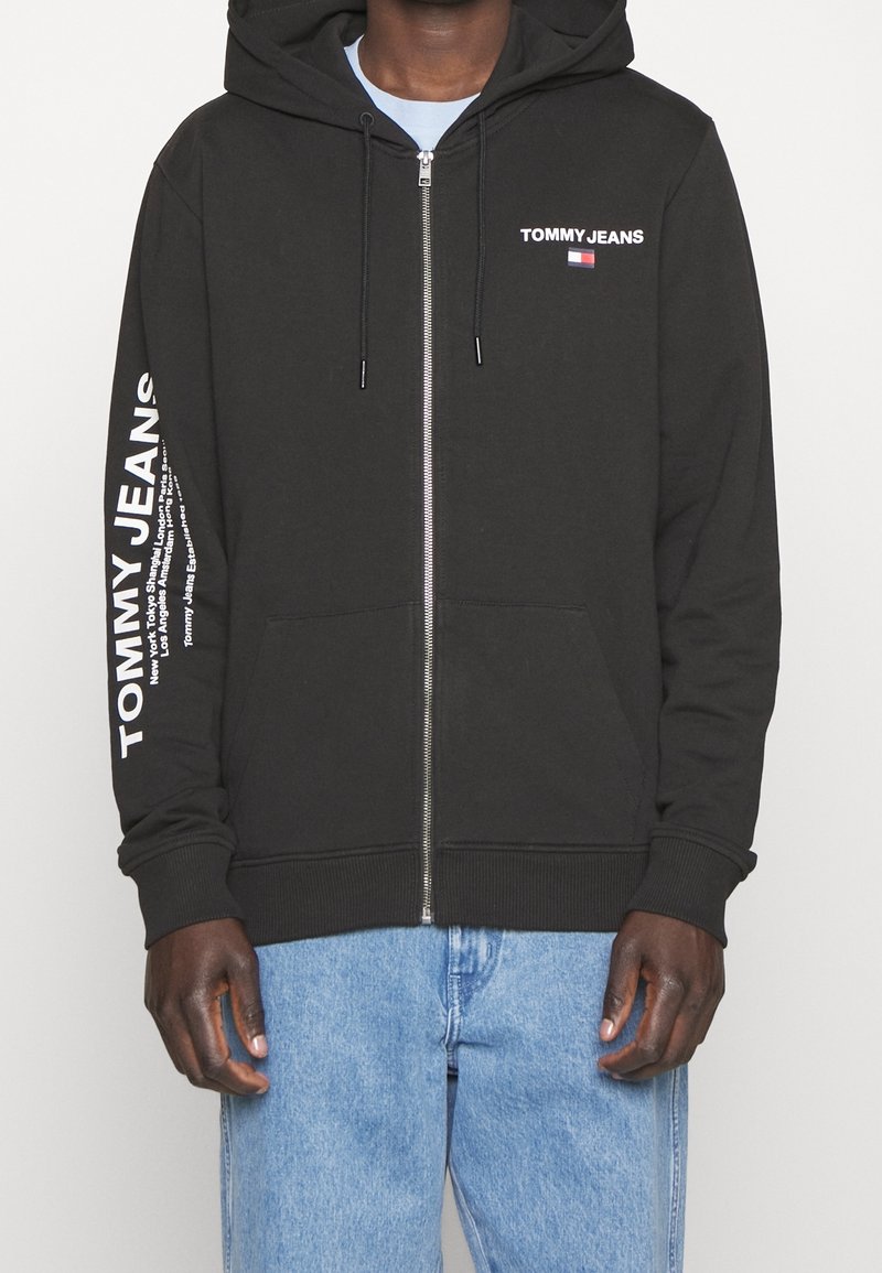 Sudadera con cremallera negra, con bolsillos frontales y capucha ajustable. Presenta el texto y el logo en blanco "TOMMY JEANS" en el pecho y en la manga.