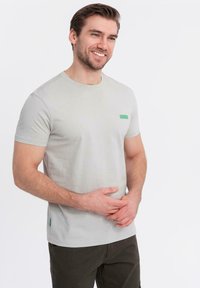 T-shirt a maniche corte grigio chiaro in cotone, con un piccolo etichetta verde sul lato sinistro del petto e un accento verde sull'orlo laterale.