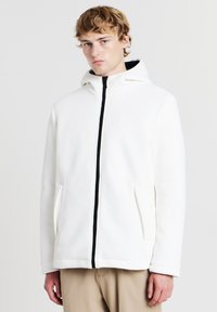 Weiße Kapuzenjacke mit geribbelter Textur, schwarzem Reißverschluss, zwei Seitentaschen und strukturierter Passform. Entworfen für einen modernen, minimalistischen Look.