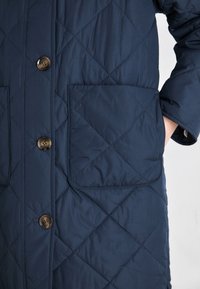 Veste matelassée bleu marine avec un devant boutonné, deux poches plaquées et une marque discrète. Elle présente un tissu doux et lisse ainsi qu un col structuré.