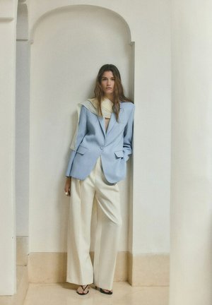 Femme debout dans une alcôve blanche portant un blazer bleu clair, un pantalon blanc à jambes larges, des sandales noires et une écharpe crème drapée.