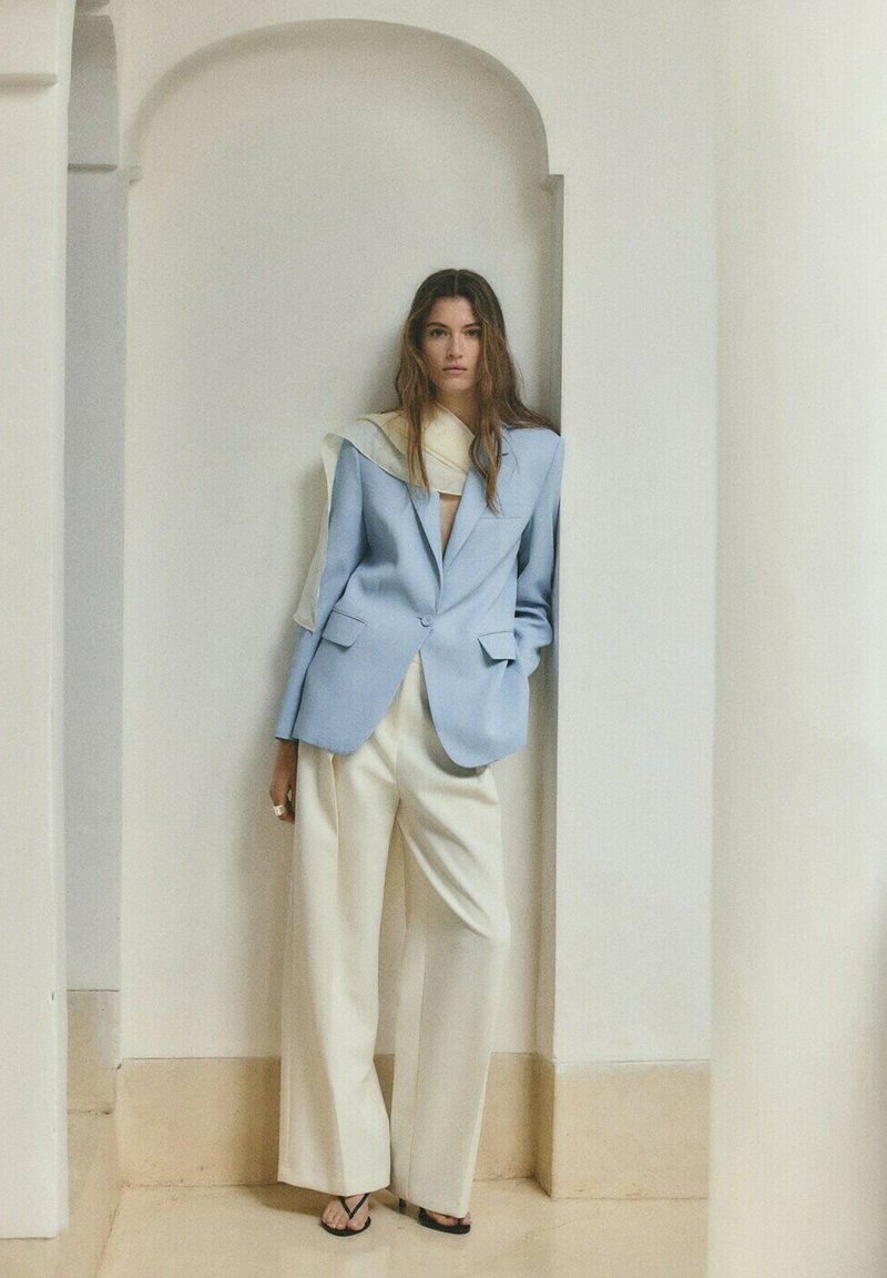 Femme debout dans une alcôve blanche portant un blazer bleu clair, un pantalon blanc à jambes larges, des sandales noires et une écharpe crème drapée.