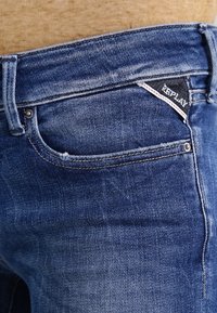 Vaqueros de mezclilla azul con un acabado desgastado, que presentan una pequeña etiqueta negra "Replay" y un diseño clásico de cinco bolsillos.