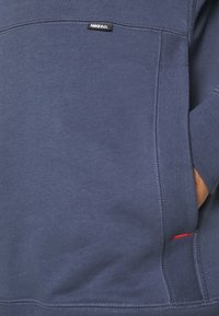 Sweat-shirt bleu marine en tissu doux avec un motif cousu, comprenant une étiquette de logo et un accent rouge sur la poche latérale.