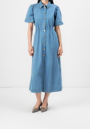Kobieta ubrana w niebieską, midi sukienkę dżinsową na guziki z krótkimi bufkami rękawów oraz szare szpilki z noskiem w szpic, stojąca na białym tle.