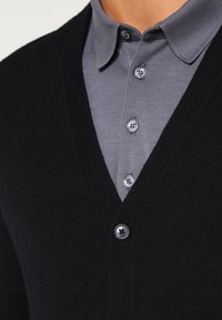 Černý kardigan s výstřihem do V vrstvený přes šedou košili s límečkem; má texturovanou látku, knoflíky s jemnými detaily a přiléhavý střih.