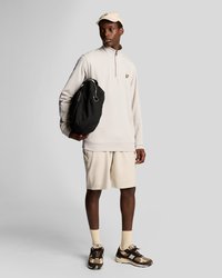 Homme portant un sweat-shirt beige à fermeture éclair quart, un short assorti et une casquette, tenant un sac à dos noir, se tenant devant un fond gris uni.