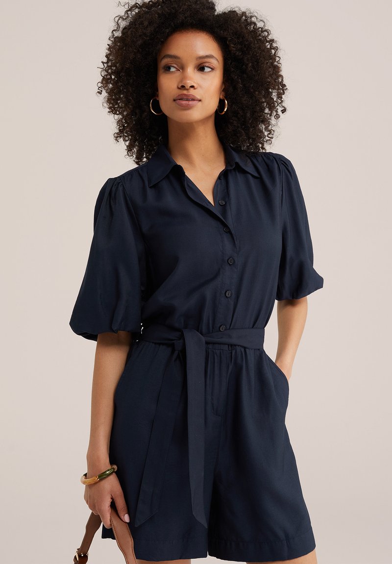 Femme aux cheveux bouclés portant un combishort bleu marine ceinturé avec des manches bouffantes, tenant une bandoulière de sac marron, regardant sur le côté.
