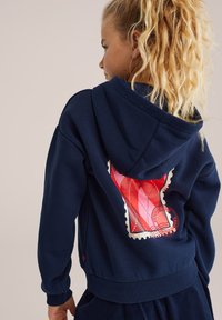 Jūras zilā kapucīna krekls, kas aizmugurē rotāts ar spilgti sarkanu un baltu grafisko dizainu, ar šuvju detaļām un ribotām piedurknēm.