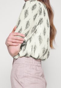 Blouse vert clair avec des motifs de feuilles noires, manches courtes à volants, et ourlet froncé, associée à un pantalon texturé de couleur rose pâle.