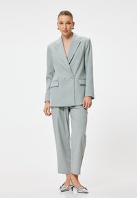 Costume ajusté gris clair avec une veste à double bouton et un pantalon à jambes droites, présentant une texture subtile et des poignets contrastants. Chaussures argentées.
