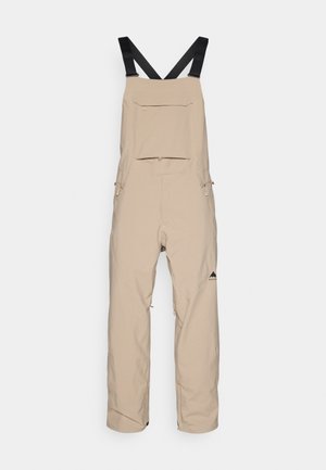 Salopette beige avec des bretelles noires ajustables, dotée d'une poche avant et de zips sur les côtés. Fabriquée en matériau durable et résistant aux intempéries.