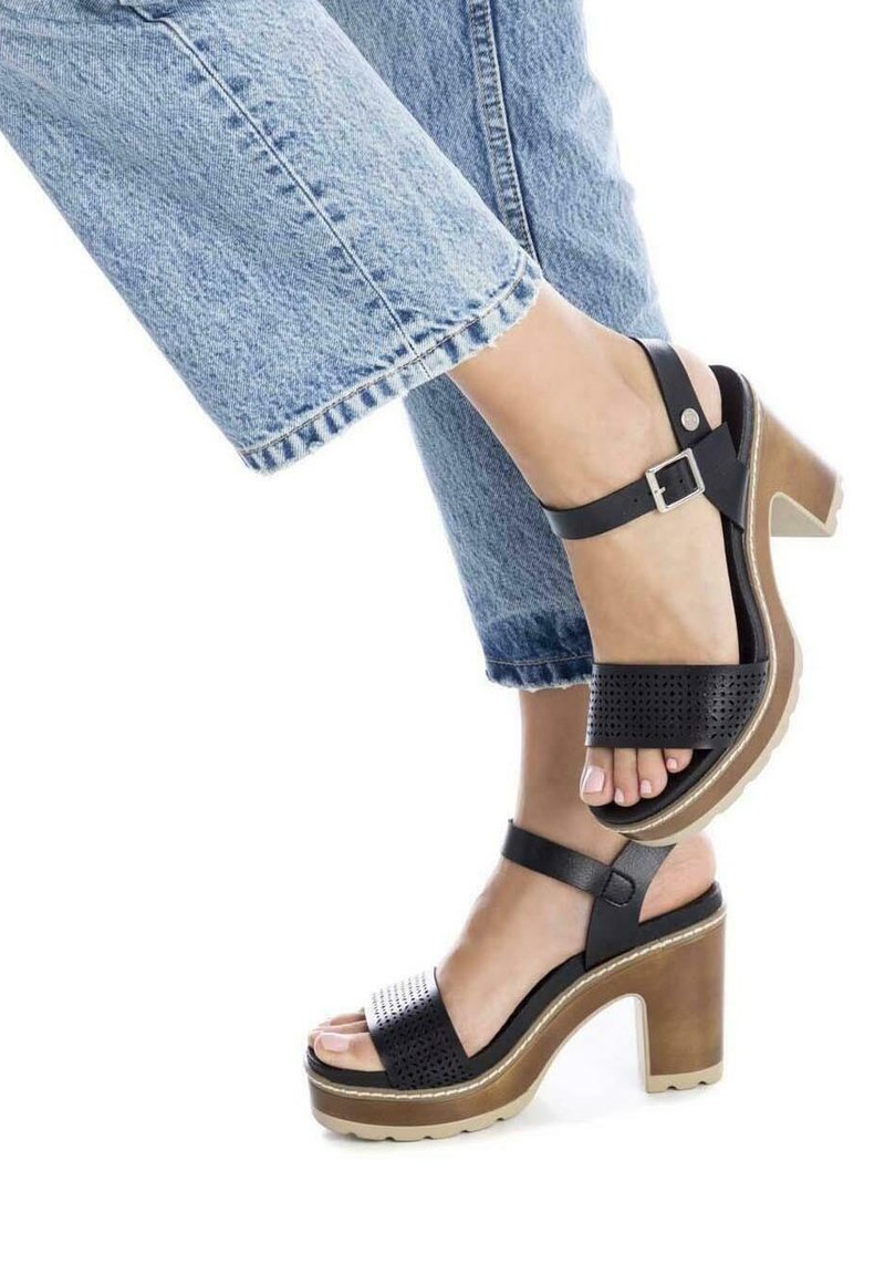 Sandalias de plataforma de cuero negro con una correa perforada, hebilla ajustable en el tobillo y tacón cuadrado. Combinadas con unos vaqueros cortos de mezclilla azul.