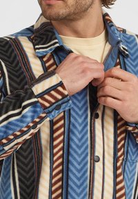 Hombre vistiendo una camisa de forro polar a rayas azules, marrones, crema y negras, abrochándola sobre una camiseta de cuello redondo beige.