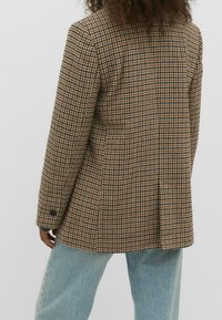 Blazer marron à motif pied-de-poule avec un design croisé, des revers crantés et des poignets boutonnés, assorti à un jean large bleu clair.