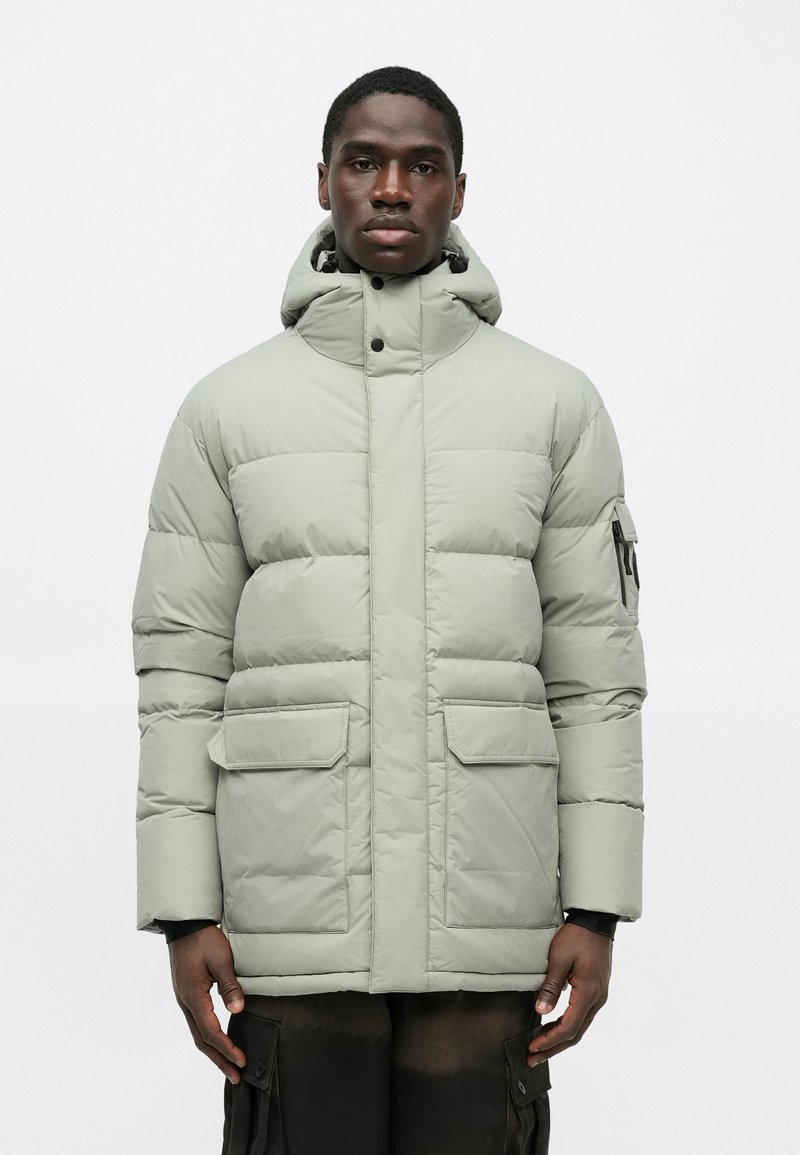 Polstret vinterjakke i lys grå, med høj krave, lynlås lukning, to frontlommer og en aftagelig hætte. Glat tekstur.