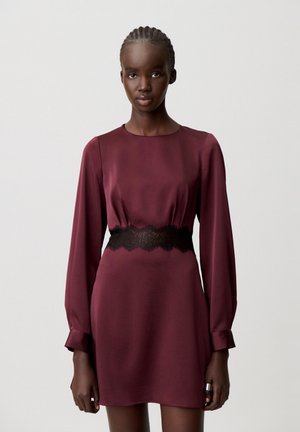 Femme aux cheveux tressés portant une robe bordeaux à manches longues avec un détail en dentelle noire à la taille, debout devant un fond uni.