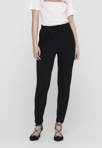 Žena v černých slim-fit kalhotách s šňůrkou v pase a černých sandálech na vysokém podpatku, doplněná bílým krátkým rukávem.