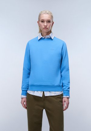 Jeune personne aux cheveux blonds courts portant un sweat-shirt bleu clair par-dessus une chemise rayée et un pantalon vert olive, debout devant un fond uni.