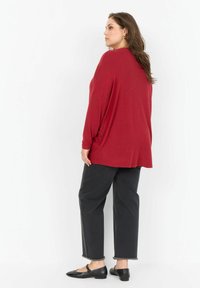 Crvena oversized pulover od mekog pletenog materijala, u kombinaciji s crnim hlačama visokog struka s raskrpljenim rubovima i crnim balerinkama.