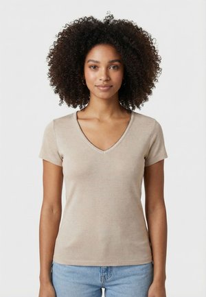 Donna con capelli ricci che indossa una maglietta beige a maniche corte con scollo a V e jeans azzurri chiari, in piedi davanti a uno sfondo bianco semplice.