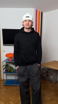 Fiatal férfi fehér sapkában, fekete kapucnis pulóverben és szürke nadrágban, egy szobában áll, amelyben TV, narancssárga lámpa, polcok és csíkos falidéző találhatók.