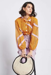 Top in rete arancione con strisce rosa, dotato di grandi bottoni e maniche a sbuffo. Il modello tiene in mano una borsa a cesta rotonda di colore crema con bordi neri.