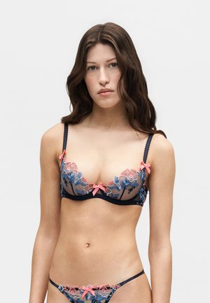 Jeune femme aux longs cheveux bruns portant une lingerie bleu marine avec une broderie florale rose et bleue et des nœuds roses, debout devant un fond uni.