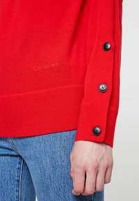 Czerwony sweter z dzianiny o zaokrąglonym dole, z czarnymi guzikami na rękawach. Dyskretne logo Calvin Klein widoczne w pobliżu dołu.