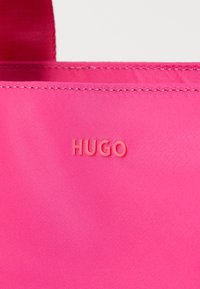 Rozā auduma soma ar izšūtiem malām, kurai ir reljefs "HUGO" logotips gaišāk rozā krāsā. Gluda tekstūra, taisnstūra forma un izturīga konstrukcija.