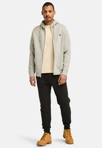 Hellgrauer Reißverschluss-Hoodie mit Kapuze und vorderen Taschen, beiges T-Shirt darunter, schwarze Hose und tanfarbene Ankle Boots mit Schnürung.