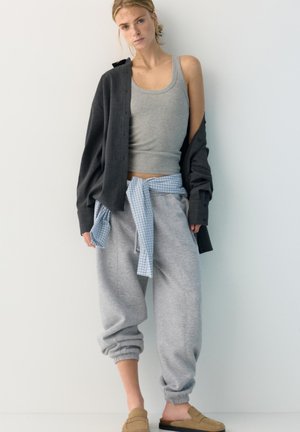 Fiatal nő réteges, hétköznapi ruhában: szürke ujjatlan felső, megkötött kockás ing, sötét nyitott ing, szürke melegítőnadrág és bézs slip-on cipő.