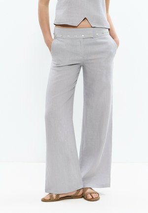 Femme portant un pantalon gris clair à jambes larges avec des boutons argentés sur la taille et des sandales marron cloutées, mains dans les poches.