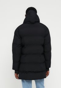 Άνδρας που φοράει μαύρο επενδυμένο χειμερινό παλτό με κουκούλα, που φαίνεται από πίσω, συνδυασμένο με ανοιχτόχρωμα μπλε τζιν, σε απλό ανοιχτό φόντο.