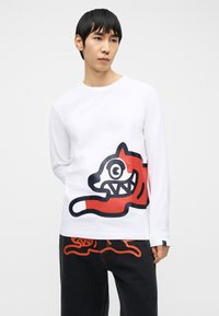 Νεαρός άνδρας που φοράει λευκό μακρυμάνικο πουκάμισο με μεγάλο κόκκινο και μαύρο καρτούν ζώο, συνδυασμένο με μαύρο παντελόνι που έχει αντίστοιχο σχέδιο.