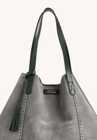 Sac fourre-tout en suède gris avec des poignées en cuir noir, présentant un détail de frange et une couture contrastante. Logo affiché à l'avant.