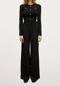 Svart stickad långärmad topp med guldbutiker och bröstfickor, tillsammans med svarta byxor med vida ben, båda sömlösa och eleganta i utseendet.