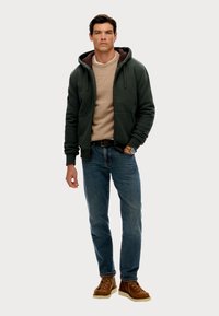 Donkergroene hoodie met een bruine binnenvoering, gedragen over een beige trui, gecombineerd met blauwe spijkerbroek en bruine veterschoenen.