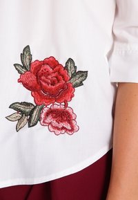 Blouse blanche ornée de roses rouges brodées avec des feuilles vertes, en tissu texturé, à manches courtes et à coupe décontractée.