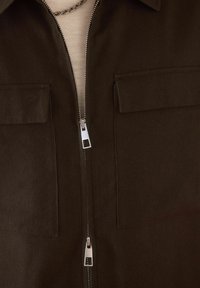 Chaqueta marrón oscuro con cremallera frontal, dos bolsillos en el pecho y una tela texturizada. Presenta detalles en cremallera plateada y un diseño con cuello.
