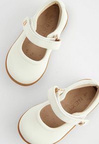 Białe skórzane buty Mary Jane z okrągłym noskiem, brązową gumową podeszwą i rzepem z złotym akcentem. Gładka tekstura i minimalistyczny design.