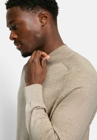 Beiger Strickpullover mit hohem Kragen, gerippten Bündchen und detaillierter Schulternaht, getragen von einer Person, die im Profil vor einem neutralen Hintergrund gezeigt wird.