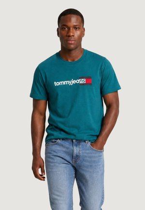 Mężczyzna w turkusowej koszulce Tommy Jeans z logo i jasnoniebieskich jeansach, stoi z jedną ręką w kieszeni na jednolitym tle.
