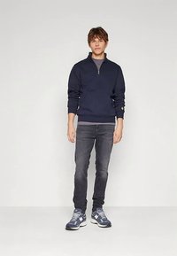 Mornarski duks s patentnim zatvaračem na vrat, u mekanom materijalu, uparan s tamnosivim trapericama i sportskim sivim tenisicama, s rebrastim rubom i manžetama.