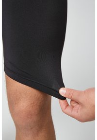 Pantaloni sportivi neri realizzati in materiale liscio ed elastico. L'orlo è aderente, con una persona che tiene il bordo, mostrando flessibilità e lunghezza.