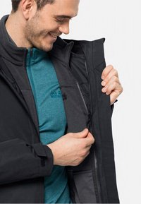 Sort jakke med lynlås foran, lagdelt over en teal fleece. Jakken har en høj krave og teksturerede stofdetaljer.