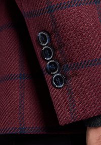 Burgundowa, teksturowana tkanina w kratkę; zawiera cztery okrągłe czarne guziki z białym brandingiem wzdłuż mankietu.