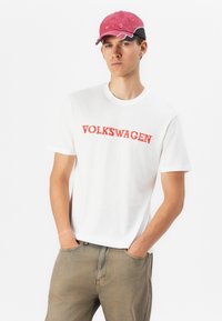 Hvid bomulds t-shirt med "VOLKSWAGEN" i fed rød skrift, parret med lyse beige jeans og en pink og sort baseballkasket.